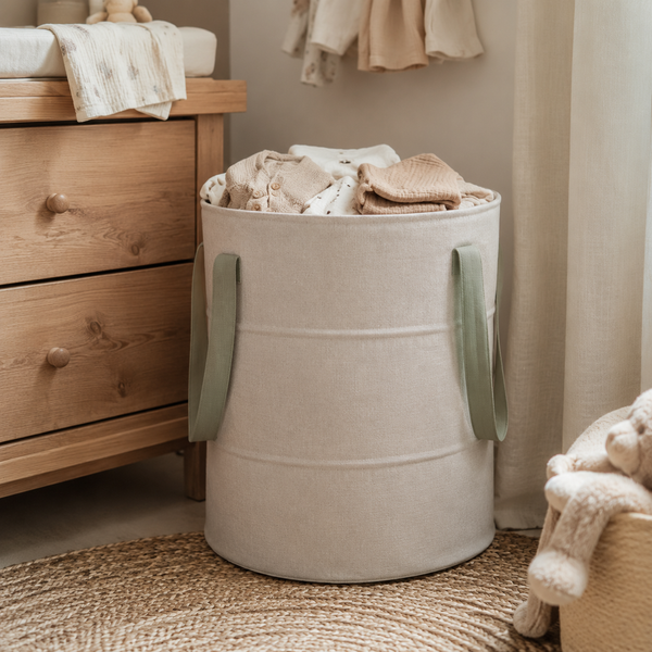 Oatmeal laundry hamper holding baby clothes beside a dresser