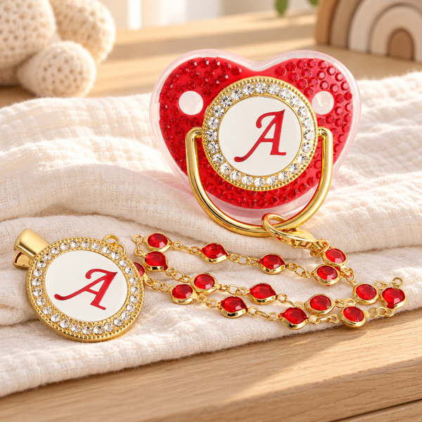 Red and gold initial A pacifier and matching clip styled on ivory muslin in a sunlit nursery